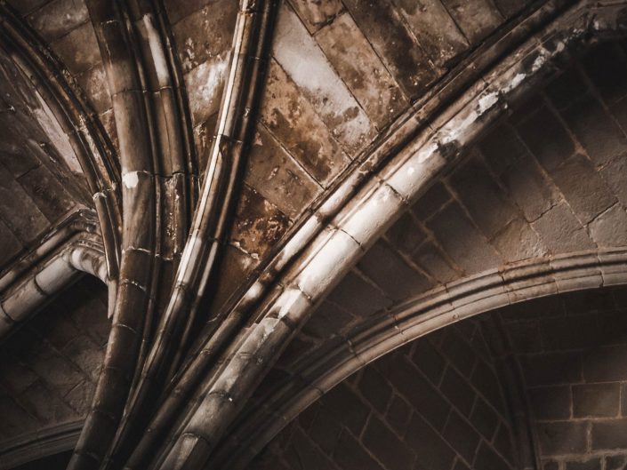 A low-angle shot of a dark, vaulted stone ceiling with intricate ribbing and arches, illuminated by soft light
