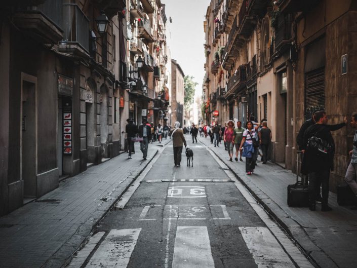 A narrow street in Barcelona with people walking on the sidewalks and a person walking a dog in the middle