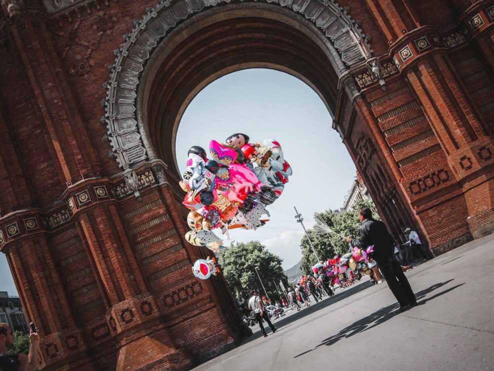 A man holding a large bunch of colorful balloons stands under a brick archway on a sunny day in Barcelona