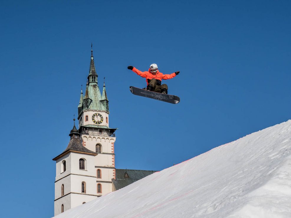 A snowboarder in an orange jacket mid-air against a clear blue sky, with a historic clock tower in the background