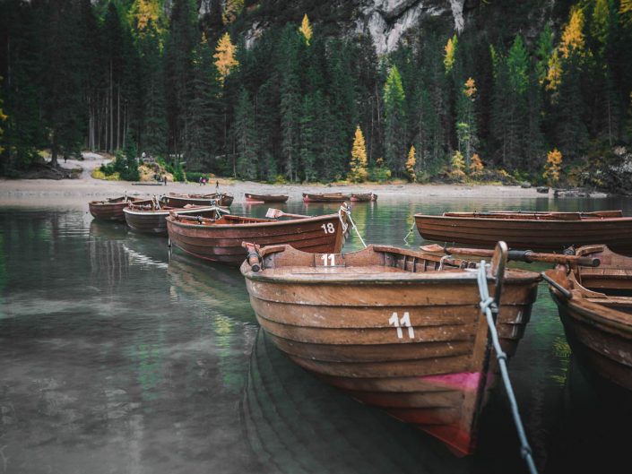 Wooden rowboats with numbers on their sides floating on a clear lake surrounded by a forest with autumn-colored trees