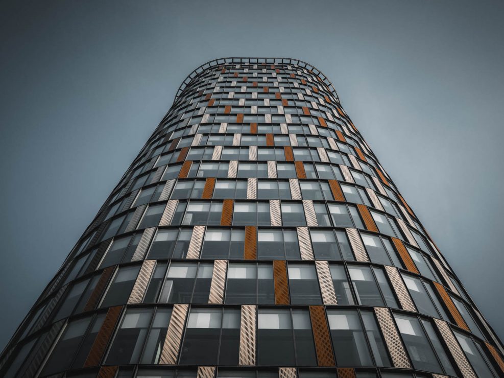 A low-angle shot of the curved facade of the Bratislava Twin City building, featuring a pattern of dark windows and alternating brown and white textured panels under a muted sky