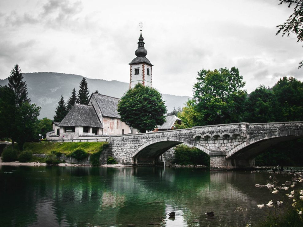 Stone bridge over a lake with a church and mountains in the background under a cloudy sky