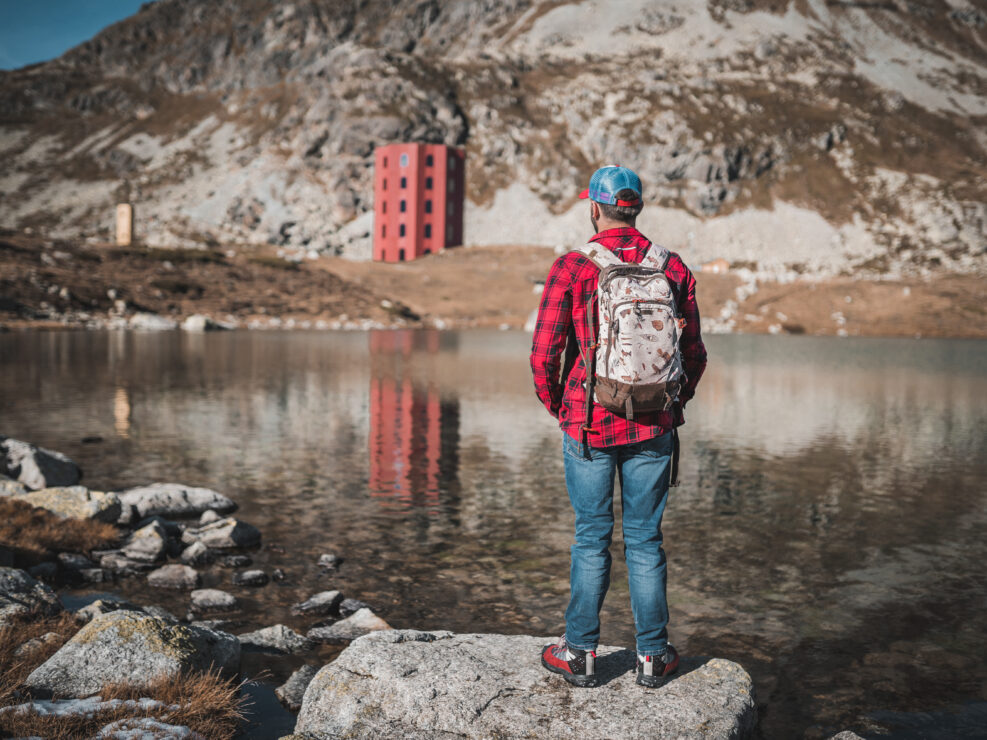 A man in a red plaid shirt, cap and backpack stands on a rock by an alpine lake, looking at a red tower below a rocky mountain