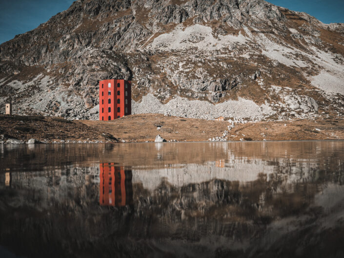 A red tower at the foot of a rocky mountain, reflected in the still water of an alpine lake