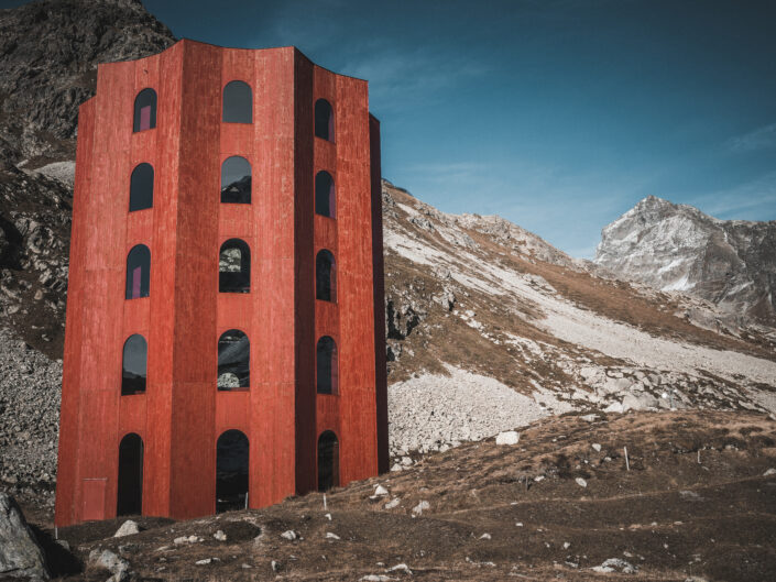 A tall red wooden tower with rows of arched windows on a rocky alpine slope, with a snow-dusted peak under a blue sky