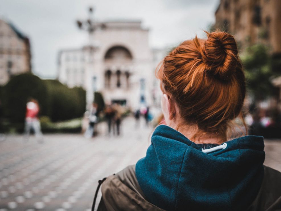 A woman with red hair in a bun, seen from behind, looking at a blurred city square with a large building