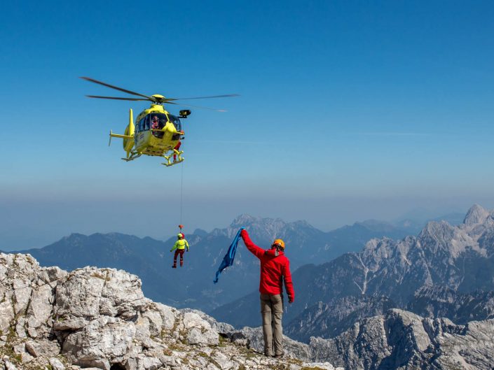 A yellow helicopter hovers over a mountain peak, lowering a person on a rope while another person waves a blue flag