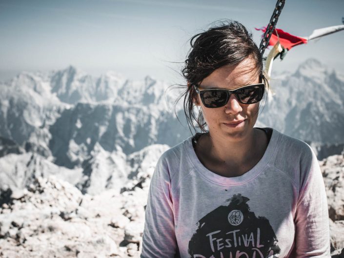 A woman in sunglasses and a light pink sweatshirt with dark hair pulled back, standing on a rocky mountain peak with a vast mountain range in the background