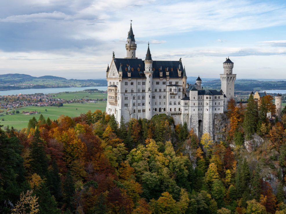 Neuschwanstein Castle, a white fairytale-like castle with many towers, sits atop a hill surrounded by colorful autumn trees
