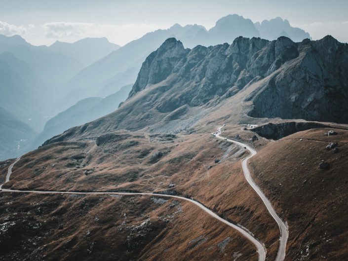 A winding road snakes through the brown, grassy slopes of the Julian Alps in Slovenia, with hazy mountains in the background