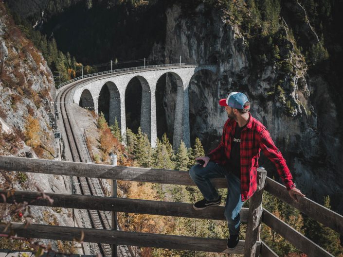 A man in a red plaid shirt and baseball cap sits on a wooden fence overlooking the Landwasser Viaduct