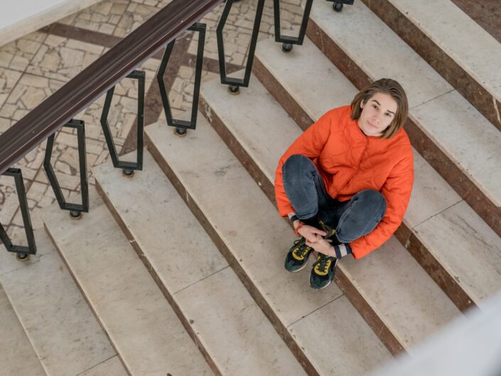 Andrea Kutlíková in an orange jacket sitting on marble stairs with a wrought-iron railing, seen from above