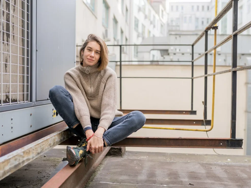Andrea Kutlíková in a beige knit sweater sitting on a steel beam on a city rooftop, smiling at the camera