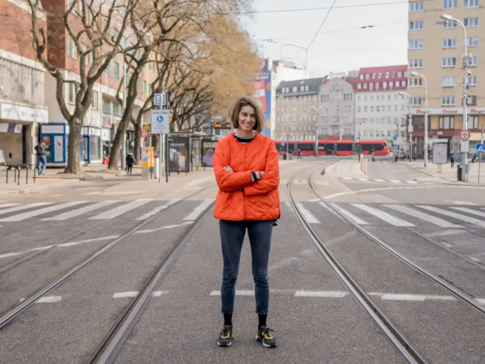 Andrea Kutlíková in an orange quilted jacket standing with crossed arms between tram tracks at Námestie SNP in Bratislava