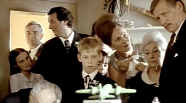Film still of a formally dressed family crowded into a small room, a boy in a suit looking at a green toy aeroplane