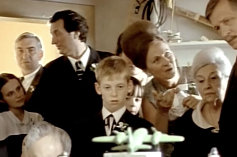 Film still of a formally dressed family crowded into a small room, a boy in a suit looking at a green toy aeroplane